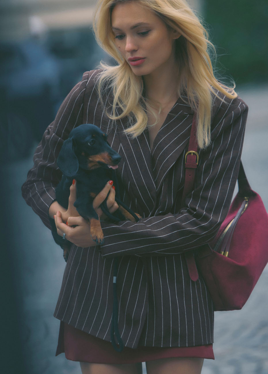 Blonde woman in black pinstripe blazer holding a black dachshund puppy, wearing a burgundy shoulder bag outdoors.