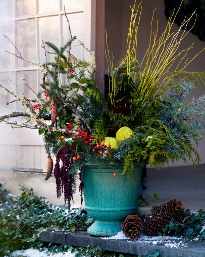 Festive winter arrangement in turquoise urn with evergreen branches, red berries, yellow twigs, and pine cones on snowy doorstep.