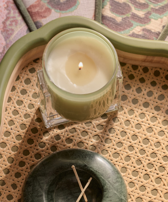 Lit green candle in glass jar on woven rattan tray with green ceramic dish containing incense sticks below