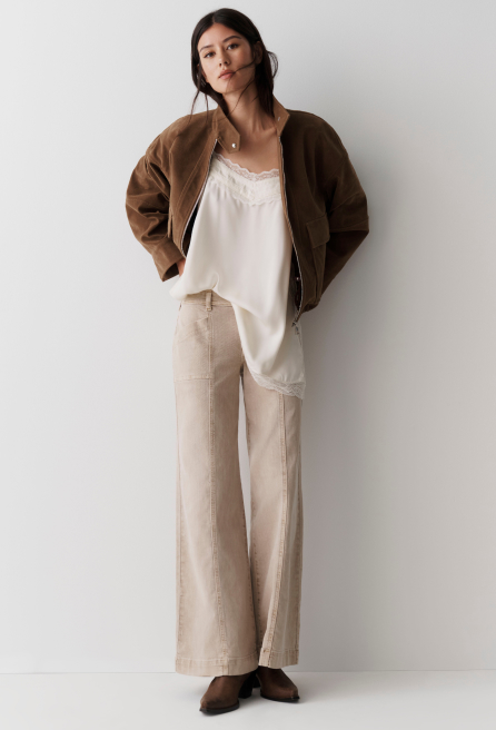 Woman wearing brown bomber jacket over white lace-trimmed camisole with beige wide-leg linen pants and brown loafers.