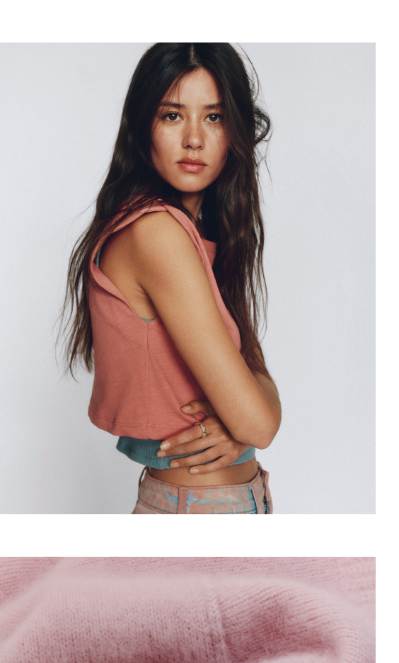 Woman with long dark hair wearing a coral pink sleeveless top and patterned pants against a white background.