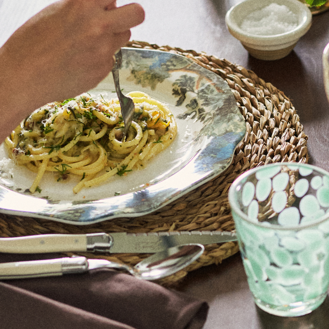 Hand twirling pasta with herbs on decorative plate over woven placemat, with mint green glass and utensils nearby.
