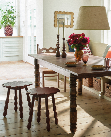 Rustic dining room with wooden table, turned leg stools, burlap lampshade, pink flowers in vase, and white storage cabinet.