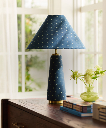 Blue polka dot table lamp with matching conical shade on wooden surface beside green vase and stacked books near window.