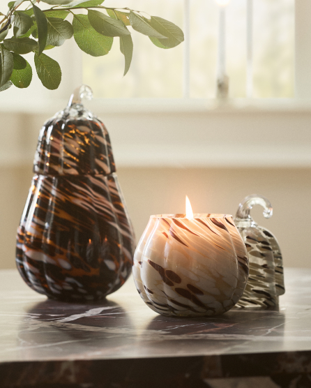 Decorative brown swirled glass pear and lit candle in marbled holder on wooden tray with green plant above