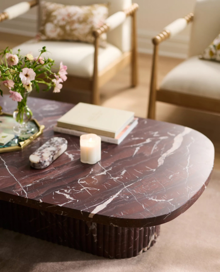 Round dark marble coffee table with white veining, topped with a candle, flowers, and decorative items, with cream upholstered chairs behind.