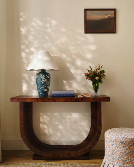 Wooden console table with blue ceramic lamp, flower vase, and books against cream wall with dappled sunlight and framed artwork.