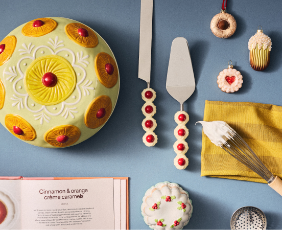 Yellow decorated cake with pineapple pattern, cookies, utensils, and recipe book on blue surface with yellow napkin.