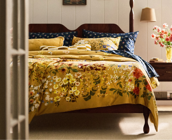 Wooden bed with golden yellow floral bedding and navy blue pillows in a bright bedroom with white walls and a bedside lamp