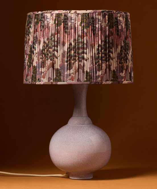 Table lamp with round pale pink ceramic base and pleated floral shade in pink, green, and cream tones on brown background.
