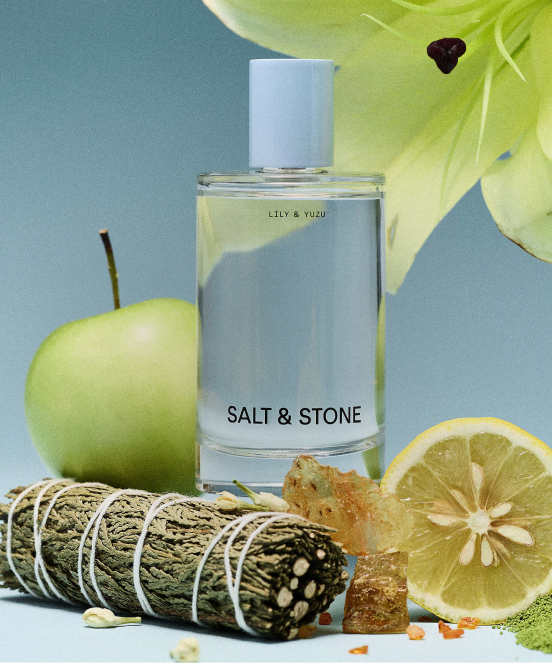 Clear perfume bottle labeled SALT & STONE LILY & YUZU surrounded by green apple, lemon slice, sage bundle, and crystals on blue surface
