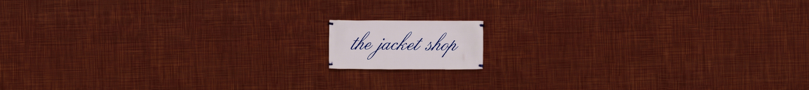 White rectangular sign with blue cursive text reading "the jacket shop" on a dark brown woven textured background