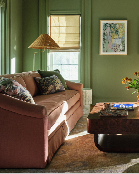 Green-walled living room with pink velvet sofa, wicker floor lamp, framed artwork, and dark wood coffee table with orange flowers.