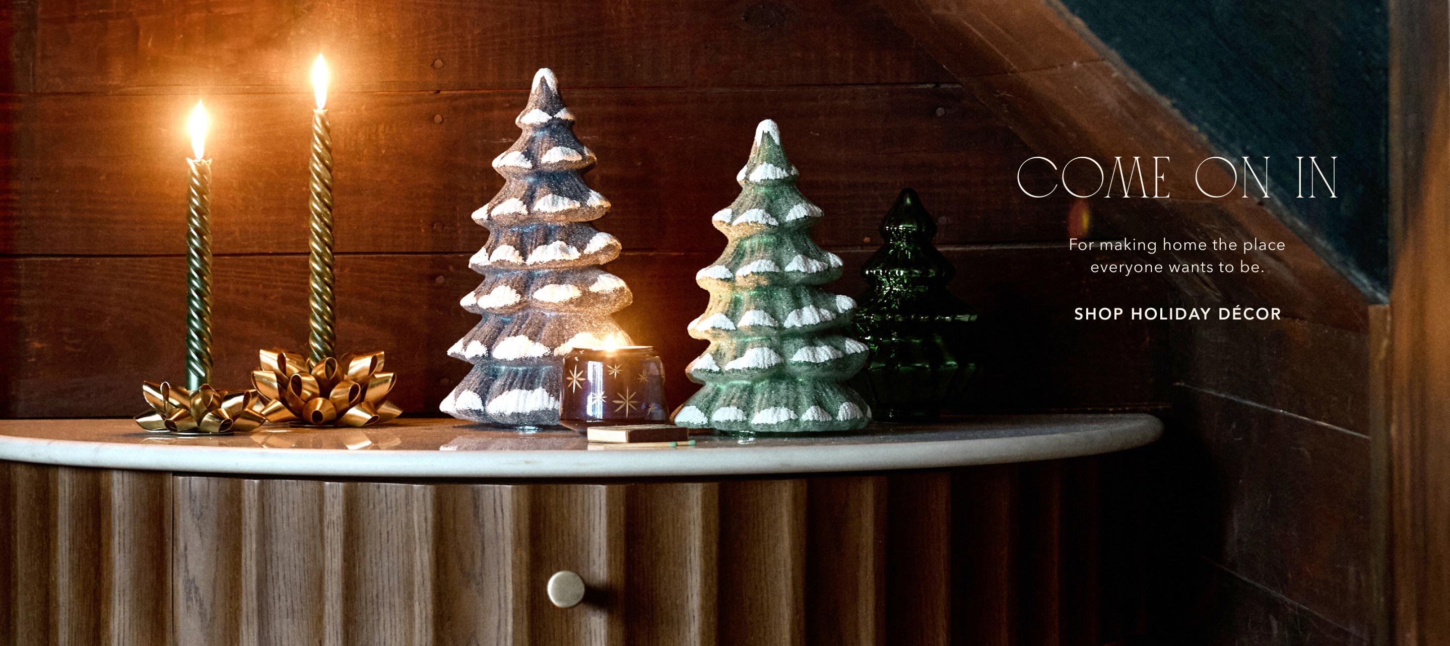 Holiday decor display with snow-dusted ceramic trees, lit candles, and text reading COME ON IN and SHOP HOLIDAY DÉCOR.