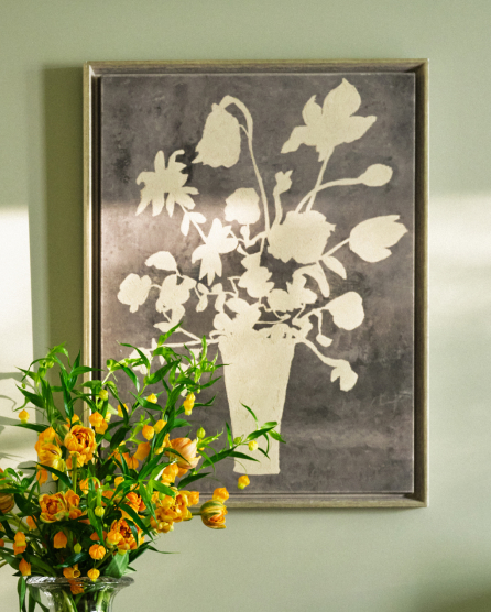 Framed floral artwork with white silhouette of flowers in vase on gray background, displayed above orange and yellow roses on green wall.