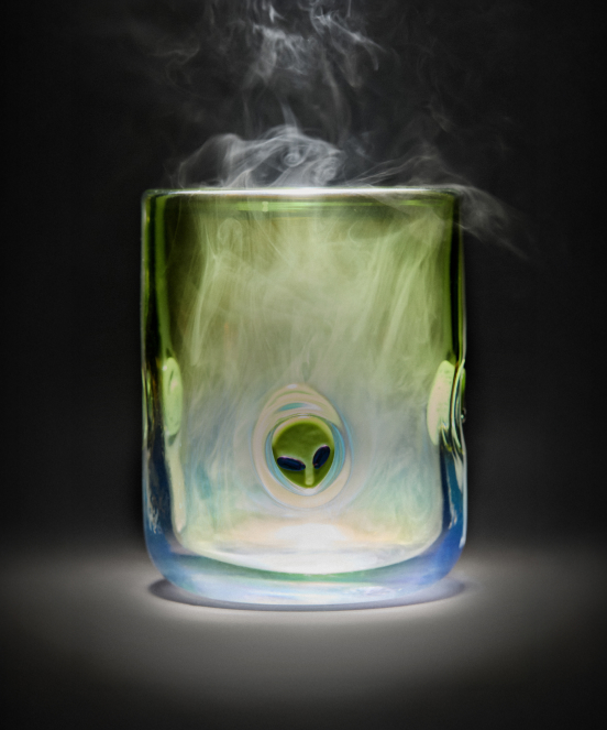 Translucent green and blue glass with alien face design, steam rising from top against dark background.