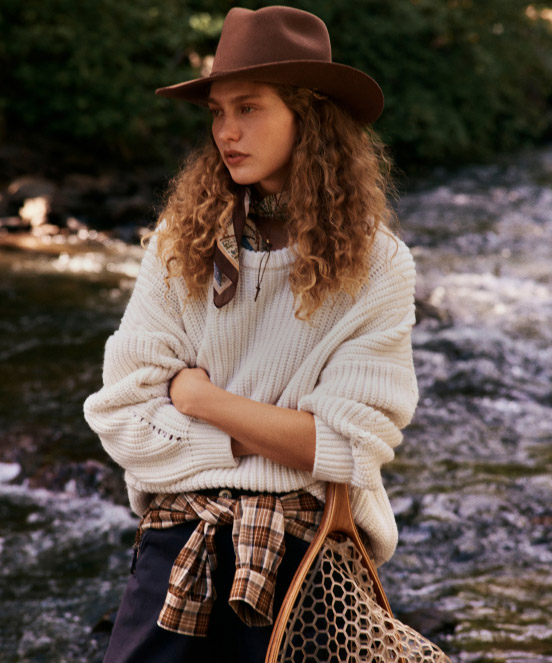 Woman in brown fedora hat and cream sweater with patterned scarf by a river, holding a honeycomb-patterned bag.
