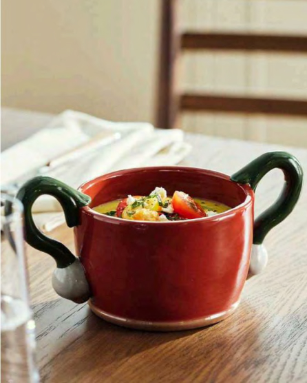Red ceramic bowl with green handles filled with colorful vegetable soup on a wooden table with white napkin nearby.