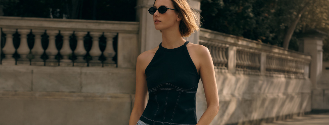 Woman in black halter top and sunglasses standing outdoors near stone balustrade with trees in background.