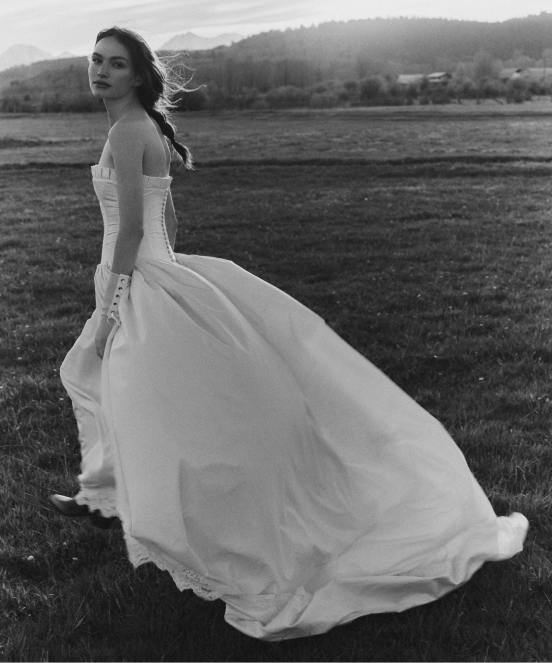 Black and white photo of a bride in a flowing white wedding gown standing in a field with hills in the background.