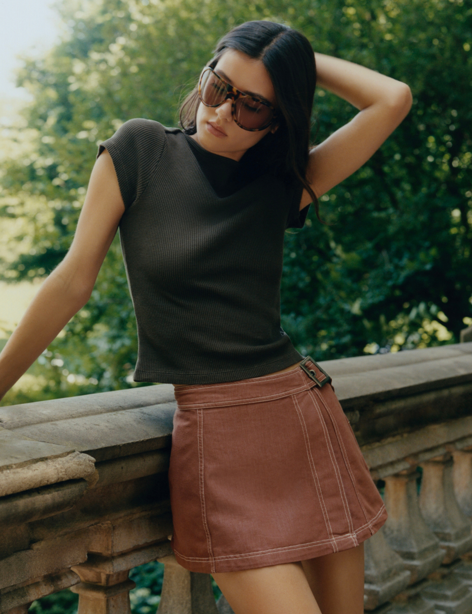 Woman in tortoiseshell sunglasses, black sleeveless top, and brown mini skirt posing outdoors with green foliage background.