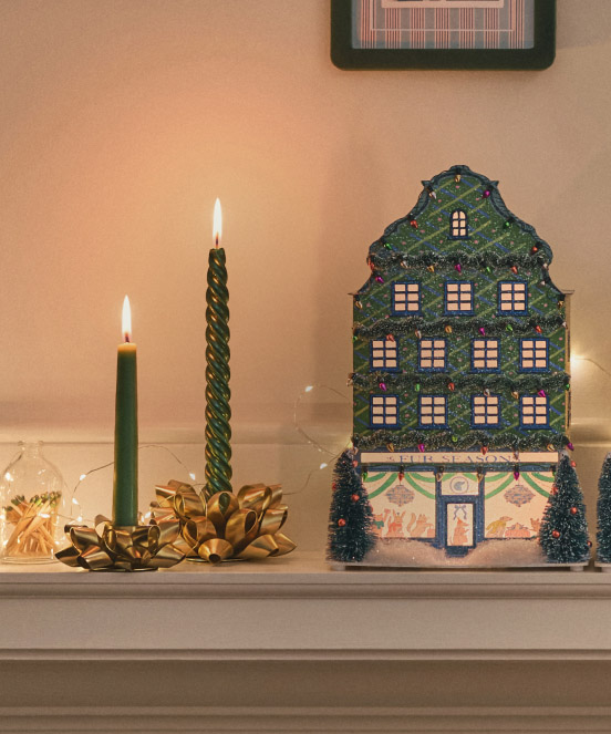 Decorative lit Christmas house with illuminated windows next to two green candles and gold bow on white mantel.