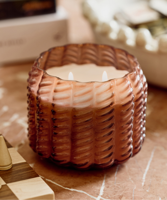 Amber-colored basket-weave textured candle with two wicks on a wooden surface with decorative items in background.