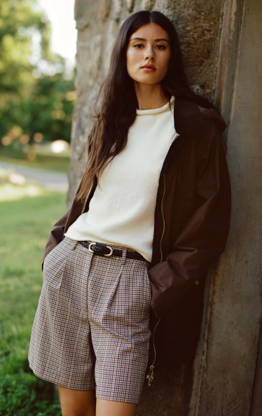 Woman with long dark hair leaning against stone column wearing brown coat, white sweater, and plaid shorts outdoors.
