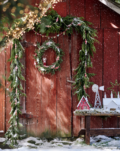Red barn doors decorated with evergreen garland spelling JOY, wreath, snow, and miniature village display with twinkling lights.
