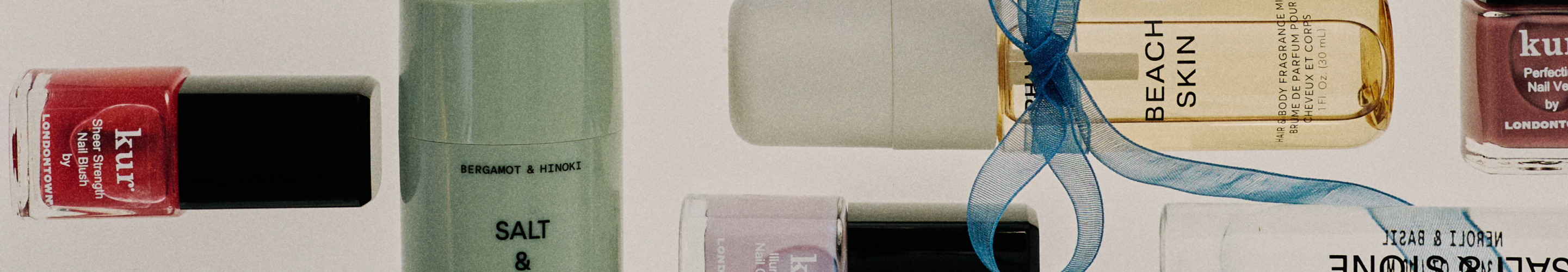 Flat lay of beauty products including nail polish bottles, a green bottle labeled "BERGAMOT & HINOKI SALT &", and "BEACH SKIN" product with blue ribbon.