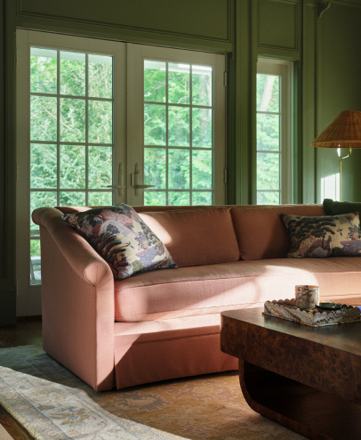 Pink curved sofa with patterned pillows in a green-walled room with large windows showing trees outside and a wooden coffee table.