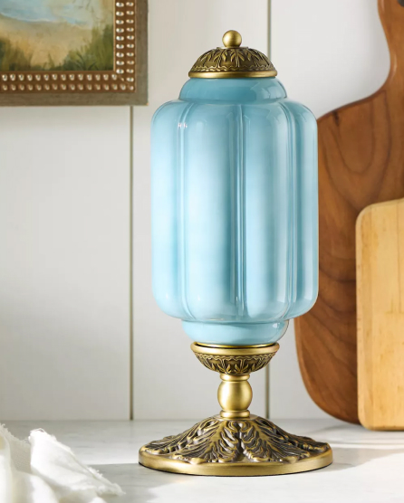 Light blue ribbed glass jar with ornate brass lid and base on white surface with framed art in background