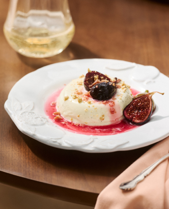 White panna cotta dessert with roasted figs and red berry sauce on a white plate, with white wine glass in background.