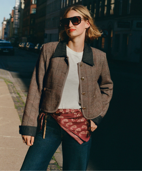 Woman in sunglasses wearing brown tweed jacket with black collar, white shirt, jeans, holding red patterned clutch outdoors.