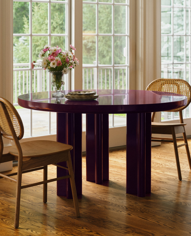 Round burgundy dining table with pink flower centerpiece and cane-back chairs near large windows with garden view.