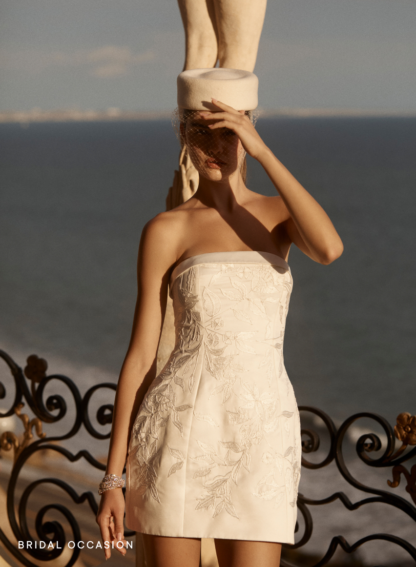 Woman in white embroidered strapless mini dress with wide-brimmed hat by the ocean, text reads BRIDAL OCCASION