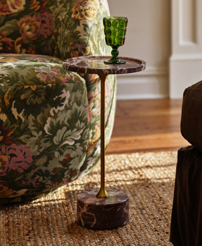 Green glass goblet on a bronze pedestal side table next to a floral upholstered chair on a jute rug