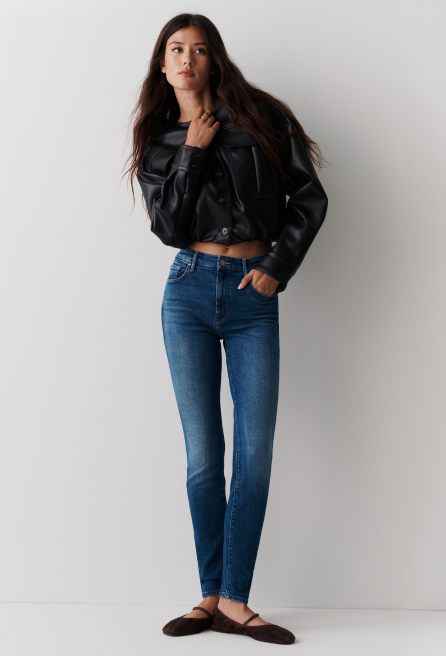 Woman wearing black cropped jacket and blue high-waisted jeans standing against white wall