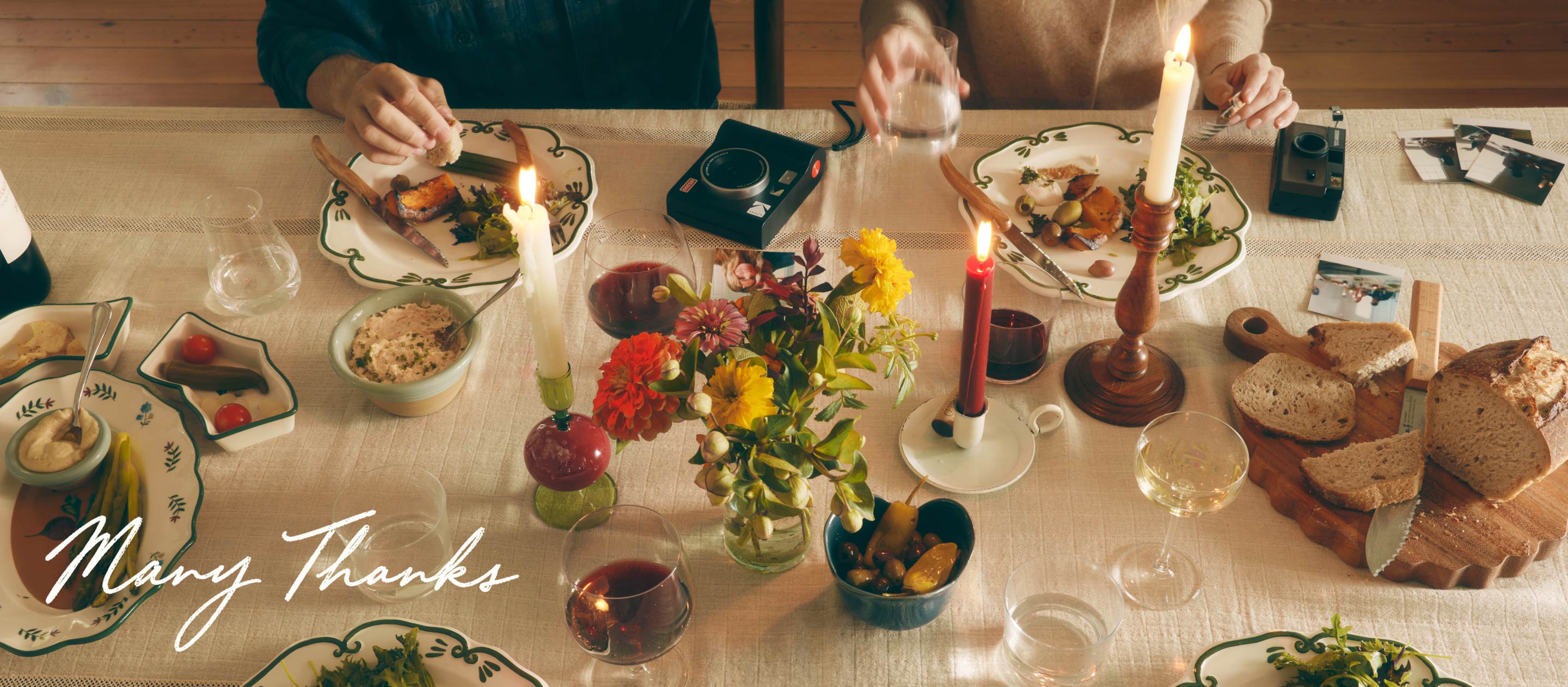 Overhead view of a dinner table with plates of food, lit candles, colorful flowers, and text reading Many Thanks.