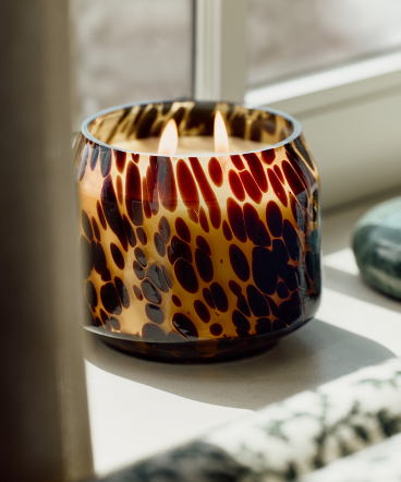 Lit candle in a decorative glass holder with amber and brown leopard print pattern on a white windowsill.