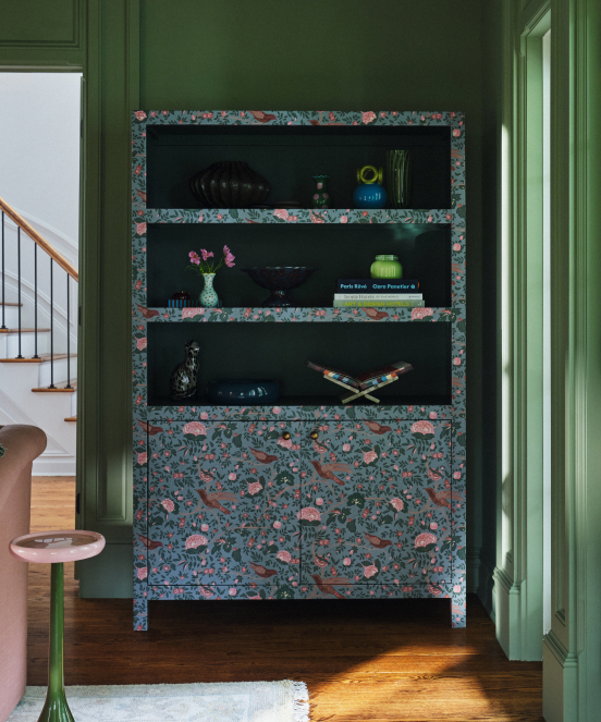 Built-in shelving unit with floral wallpaper trim against dark green walls, displaying decorative objects and books.