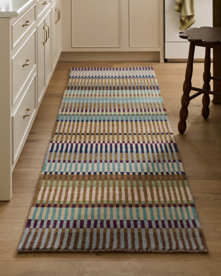 Colorful striped runner rug with blue, purple, and tan horizontal bands on hardwood floor in kitchen with white cabinets and stool.