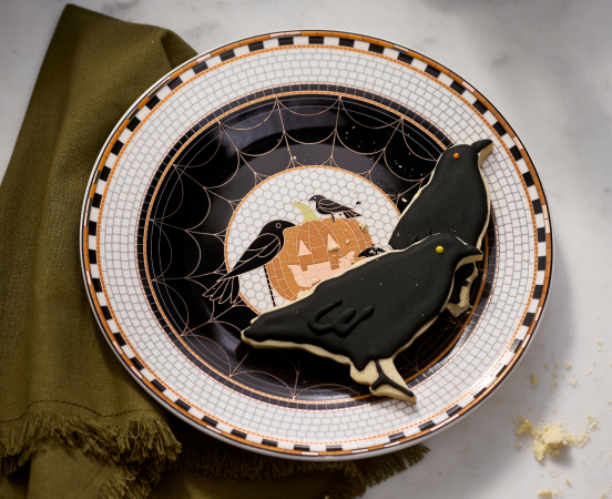 Halloween plate with black cat cookie, orange moon, ravens, and spiderweb design on white checkered rim with olive napkin.