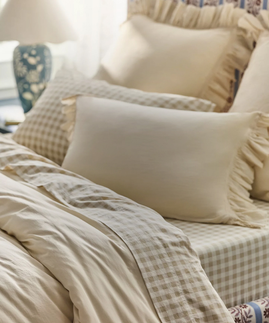 Layered bed with cream and beige gingham bedding, white pillows with ruffled edges, and a blue decorative vase in background.