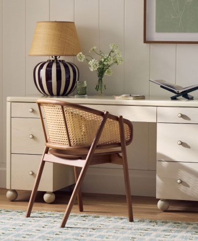 Wooden cane-back chair at white desk with striped lamp, flowers in vase, and decorative items on wood floor with patterned rug.