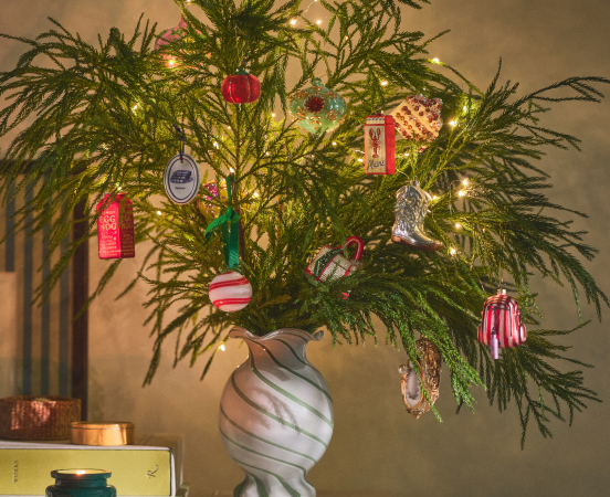 Green pine branches in a white marbled vase decorated with colorful ornaments and warm string lights against a beige wall.