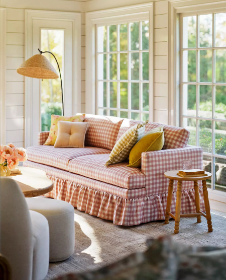 Bright sunroom with pink gingham sofa, yellow pillows, woven floor lamp, and white French doors overlooking greenery.