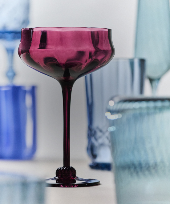 Purple coupe glass with long stem in focus, with blue and teal glassware blurred in background on white surface.