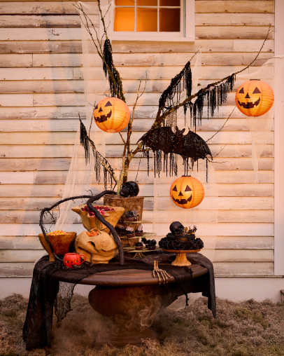 Halloween display with glowing jack-o'-lanterns hanging from bare branches, black decorations, pumpkins, and spooky items on a draped table.