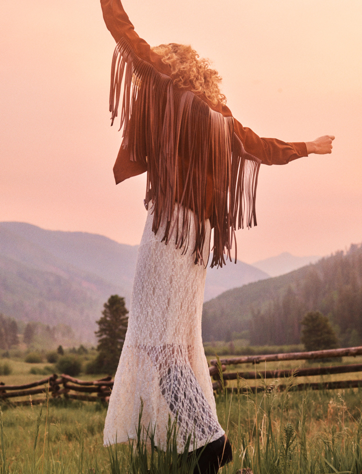 Woman with arms raised wearing brown fringed jacket and white lace skirt in mountain meadow at sunset